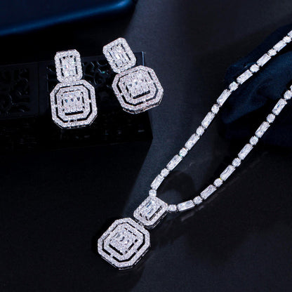 A silver-toned jewelry set featuring a necklace and matching earrings with geometric octagonal designs paved with clear stones, displayed on a dark reflective surface.